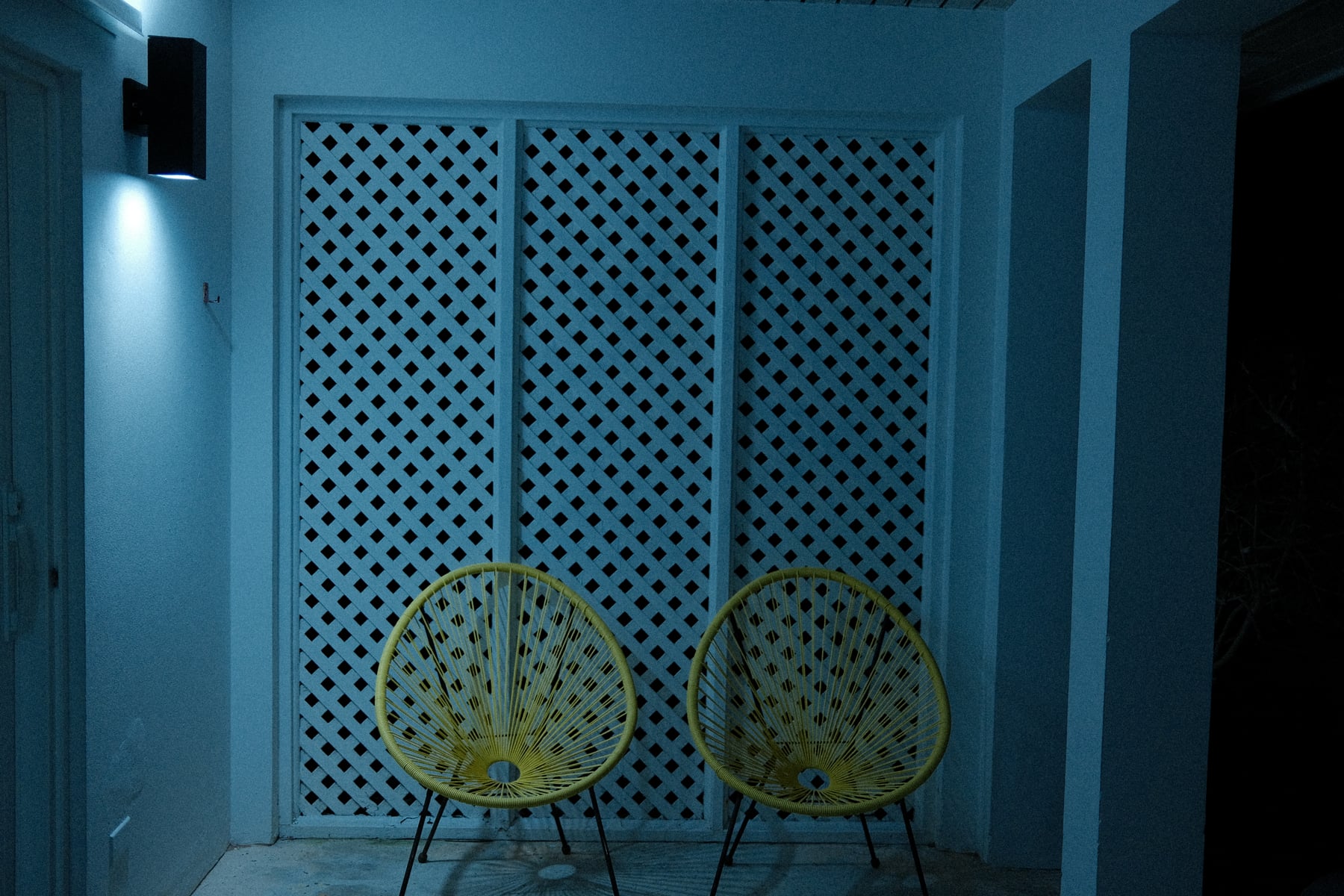 Two yellow Acapulco chairs against white diamond lattice wall at night, cool blue shadows
